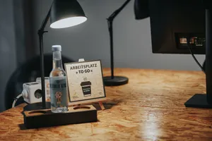 Angeschaltete Arbeitsleuchte auf einem Schreibtisch. Darunter steht eine Wasserflasche und ein Schild mit der Aufschrift "Arbeitsplatz to go". 