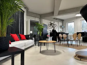 Ein offener, heller Raum im Coworking Space mit einem Sofa, mehreren Schreibtischen und Fahrrädern an der Wand. Im Hintergrund unterhalten sich zwei Frauen.