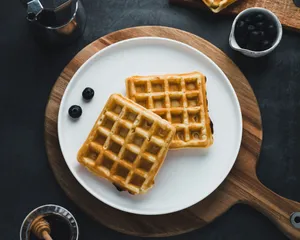 Perspektive von oben auf zwei gebackene Waffeln, die auf einem weißen Teller liegen. Der Teller steht auf einem dunklen Tisch.