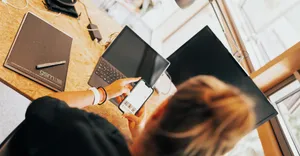 Blick über die Schulter einer Frau, die an einem Arbeitsplatz sitzt und auf ihr Handy schaut