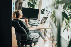 Eine Frau sitzt an einem Arbeitsplatz mit Laptop und Bildschirm und denkt nach